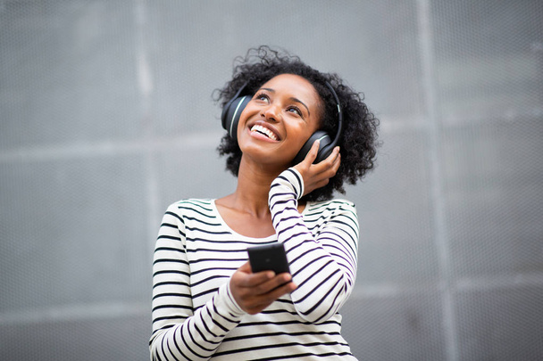 Портрет счастливая молодая черная женщина слушает музыку с сотовым телефоном и наушниками и смотрит в сторону
 - Фото, изображение