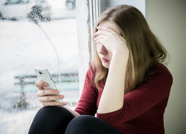 A teenager girl with long blond hair sits by the window on a windowsill with a phone in her hand and covered her face with her hand. The concept of teenage addiction to a mobile phone, the Internet, depression, isolation. - Valokuva, kuva