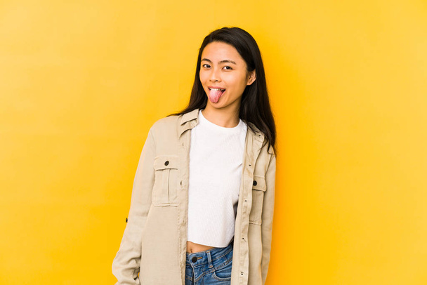 Young chinese woman isolated on a yellow background funny and friendly sticking out tongue. - Photo, Image