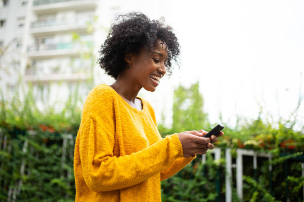 Side portret gelukkig jonge zwarte vrouw op zoek naar mobiele telefoon buiten - Foto, afbeelding