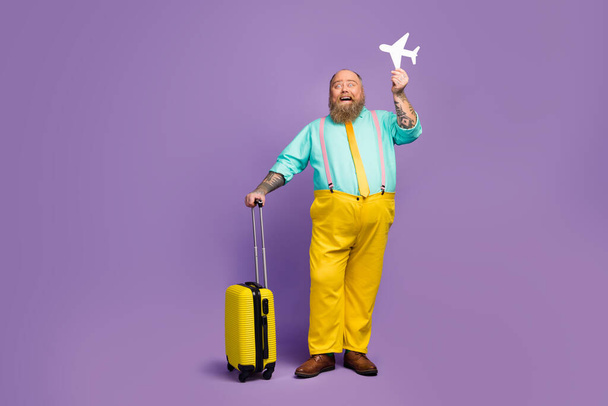 Full size photo excited man big abdomen hold papercard plane travel abroad weekend touch yellow suitcase wear shine turquoise shirt shoes pink suspenders pants isolated purple color background - Photo, Image