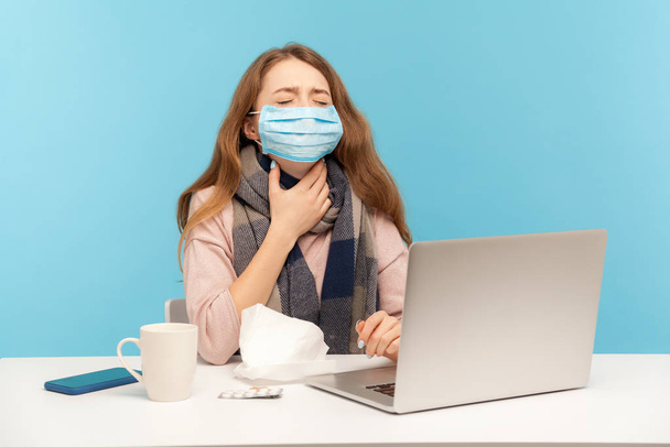Sick girl in hygienic face mask sitting at desk with laptop, holding painful neck suffering sore throat, ill freelance woman with flu working from home office. indoor studio shot, blue background - Photo, image