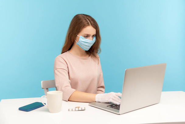 Office worker wearing facial mask and hygienic gloves while typing on laptop, protecting her hands and face to prevent contagious disease, viral infection, coronavirus 2019-ncov. indoor studio shot - Foto, afbeelding