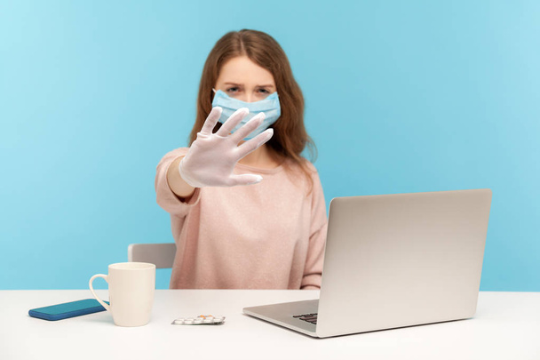 Girl in facial mask and hygienic gloves sitting at desk with laptop, showing stop, warning about danger of getting sick at office workplace, infectious disease, coronavirus epidemic. studio shot - Fotoğraf, Görsel