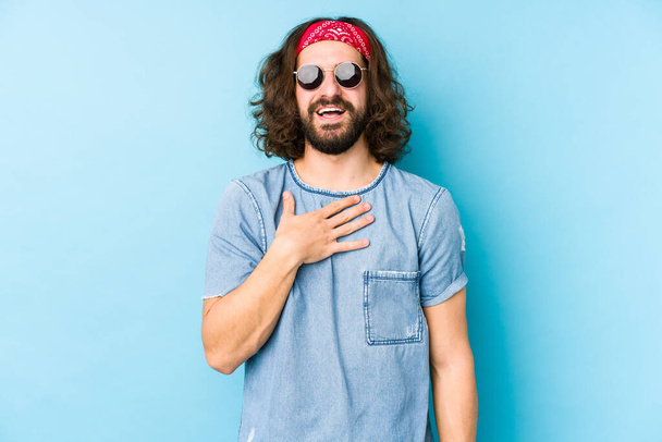 Jovem homem de cabelo longo vestindo um festival hipster olhar isolado ri em voz alta mantendo a mão no peito
. - Foto, Imagem