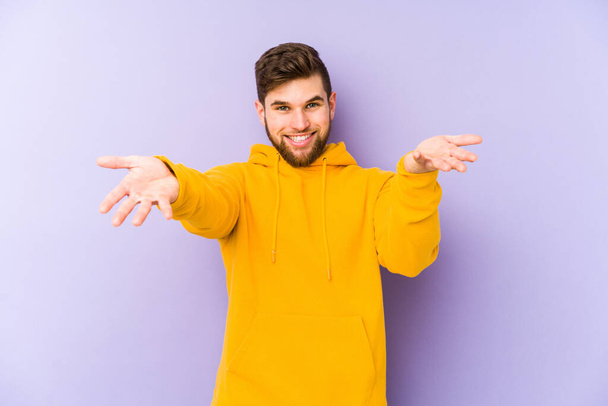 Young man isolated on purple background makes scale with arms, feels happy and confident. - Photo, Image
