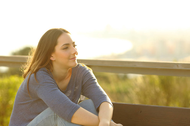 Zadowolona kobieta odwraca wzrok na ławce w parku o zachodzie słońca - Zdjęcie, obraz
