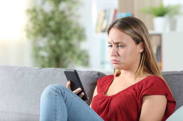 Mujer confusa después de mirar su teléfono sentado en el sofá en casa
 - Foto, imagen