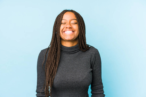 Młoda Afroamerykanka odizolowana na niebieskim tle śmieje się i zamyka oczy, czuje się zrelaksowana i szczęśliwa. - Zdjęcie, obraz