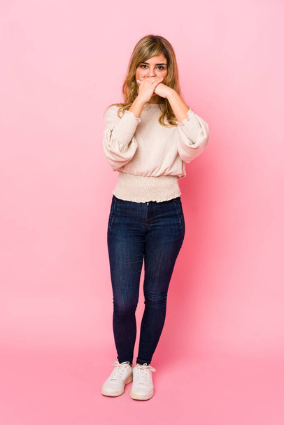 Молодая блондинка-кавказка, стоящая на розовом фоне, потрясена, закрывая рот руками
. - Фото, изображение