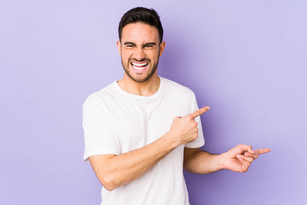 Junger kaukasischer Mann isoliert auf violettem Hintergrund, der mit den Zeigefingern auf einen Kopierraum zeigt und Begeisterung und Wunsch ausdrückt. - Foto, Bild