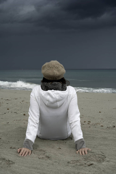 Молодая женщина сидит у моря с бурным небом, задумчиво смотрит на горизонт
 - Фото, изображение