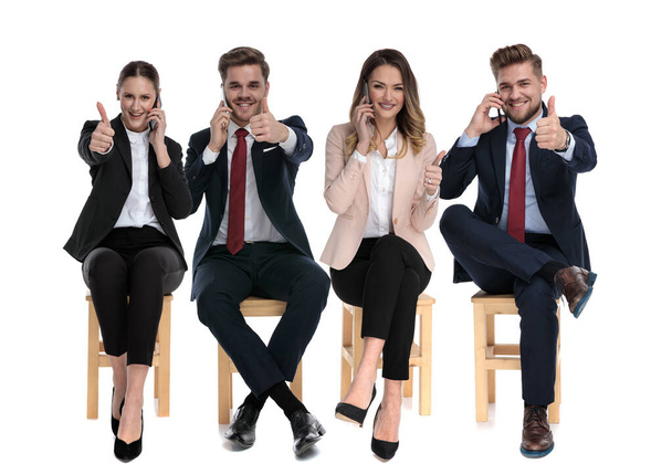 Team of 4 cheerful businessmen talking on phones and giving thumbs up while waiting for a job interview sitting on chairs on white studio background - Photo, Image