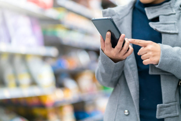 zbliżenie Azji kobieta ręka trzymać smartfon sprawdzanie ceny w supermarkecie sklep z wózek zakupy karton z rząd produktów - Zdjęcie, obraz