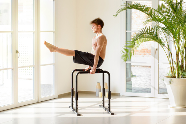 Fit mladý muž pracuje na paralelních tyčích dělat V sit cvičení během kalisthenics cvičení v moderní high key tělocvičny v konceptu zdraví a fitness - Fotografie, Obrázek