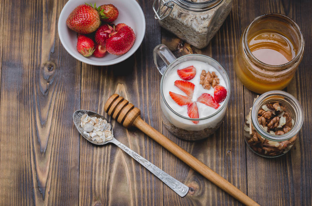 προετοιμασία γιαουρτιού με έναν κόκκο, μέλι, ξηρούς καρπούς και φράουλα σε ξύλινο τραπέζι. Άνω όψη - Φωτογραφία, εικόνα