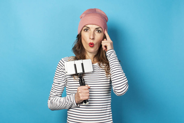 Mulher jovem vídeo blogger segura o telefone em um tripé e faz um gesto de pensar em um fundo azul. O conceito de história, vlog, selfie, blog, surpresa, choque. Banner. - Foto, Imagem