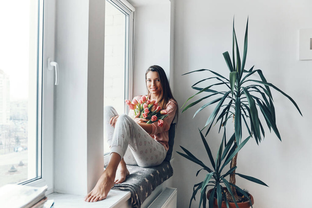 Attractive young woman in cozy pajamas enjoying her bouquet of tulips and looking at camera while resting on the window sill at home        - Фото, зображення