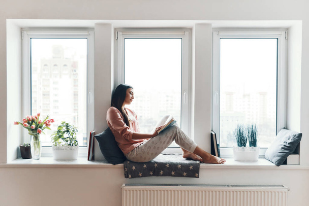 Carefree young woman in cozy pajamas reading a book while resting on the window sill at home   - Foto, Bild