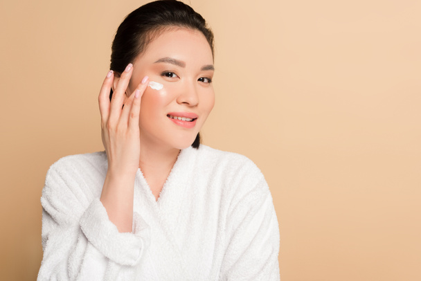 smiling beautiful asian woman in bathrobe applying face cream on beige background - Photo, Image