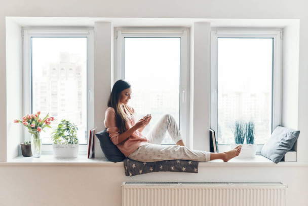 Carefree young woman in cozy pajamas using smartphone and smiling while resting on the window sill at home    - Fotografie, Obrázek