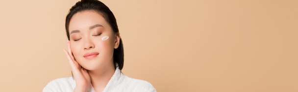 erfreut schöne asiatische Frau im Bademantel mit Gesichtscreme auf der Wange und geschlossenen Augen auf beigem Hintergrund, Panoramaaufnahme - Foto, Bild