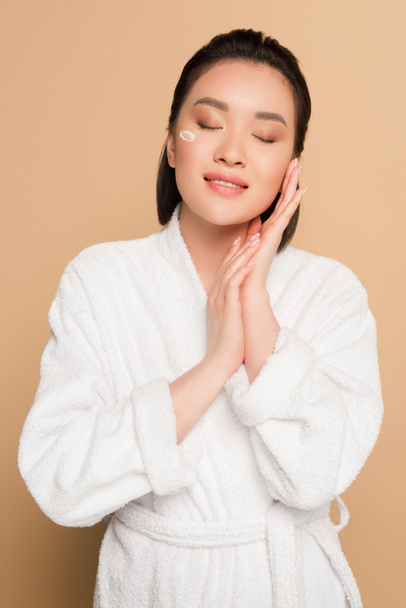 pleased beautiful asian woman in bathrobe with face cream on cheek and closed eyes on beige background - Fotoğraf, Görsel