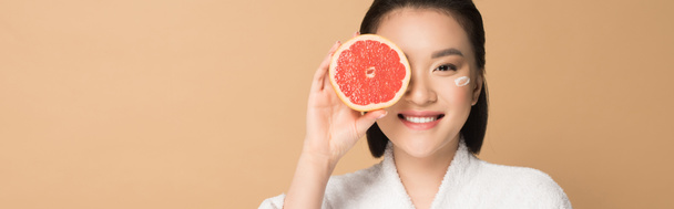 smiling beautiful asian woman in bathrobe with face cream on cheek and grapefruit half on beige background, panoramic shot - Фото, изображение