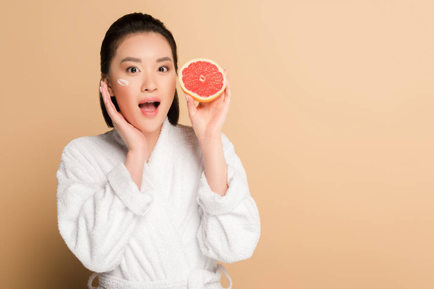 shocked beautiful asian woman in bathrobe with face cream on cheek and grapefruit half on beige background - Фото, изображение