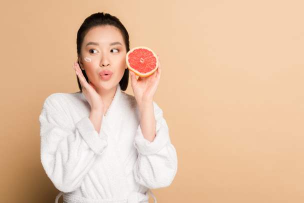 sorprendida hermosa mujer asiática en albornoz con crema facial en la mejilla y la mitad de pomelo sobre fondo beige
 - Foto, imagen