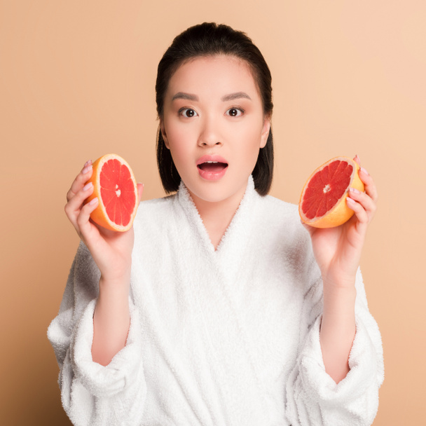 impactada hermosa mujer asiática en albornoz con mitades de pomelo sobre fondo beige
 - Foto, Imagen