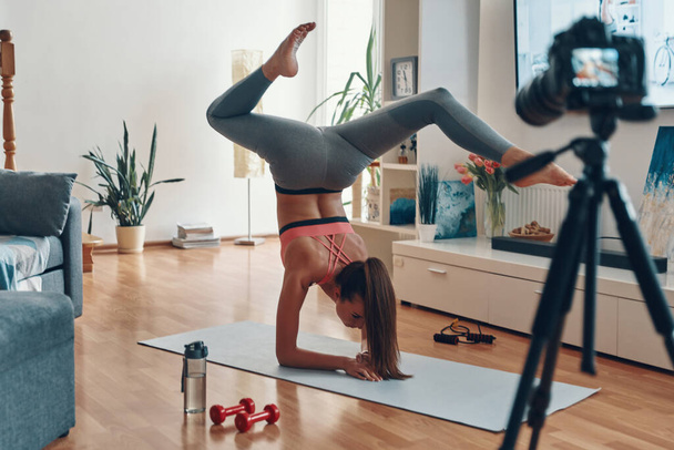Flexible young woman in sports clothing standing on hands while making social media video            - Φωτογραφία, εικόνα