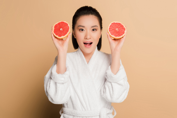 shocked beautiful asian woman in bathrobe with grapefruit halves on beige background - Photo, Image