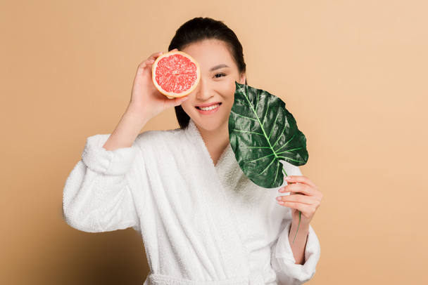 glücklich schöne asiatische Frau im Bademantel mit Grapefruit-Hälfte und Blatt auf beigem Hintergrund - Foto, Bild