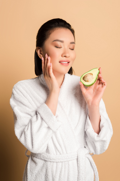 beautiful asian woman in bathrobe with avocado half touching face on beige background - Photo, Image