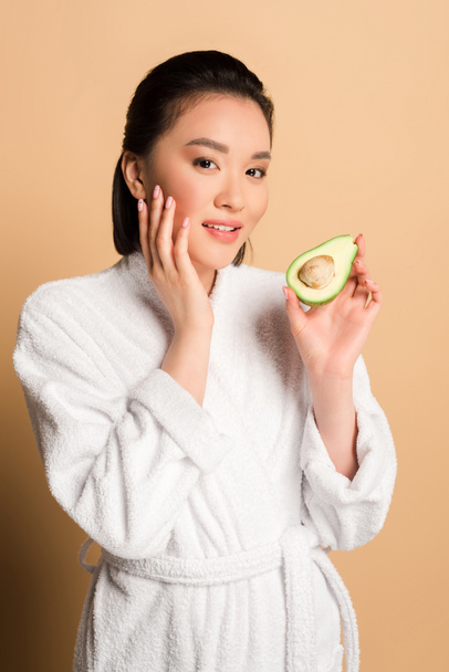 beautiful asian woman in bathrobe with avocado half touching face on beige background - Foto, Imagem