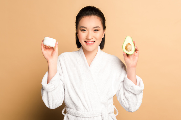 feliz hermosa mujer asiática en albornoz con aguacate mitad y crema facial sobre fondo beige
 - Foto, imagen