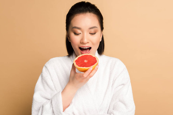 beautiful asian woman in bathrobe eating grapefruit on beige background - Photo, Image