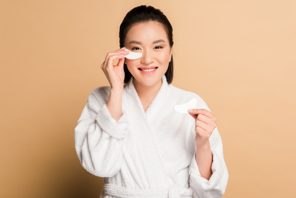 happy beautiful asian woman in bathrobe applying eye patches on face on beige background - Foto, Imagen