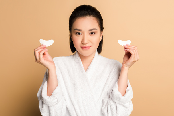 smiling beautiful asian woman in bathrobe holding eye patches on beige background - Fotografie, Obrázek