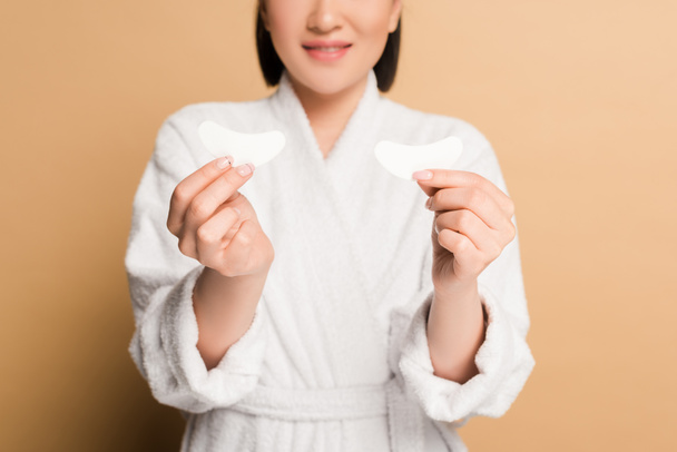 kivágott kilátás mosolygós gyönyörű ázsiai nő fürdőköpenyben gazdaság szem foltok bézs háttér - Fotó, kép
