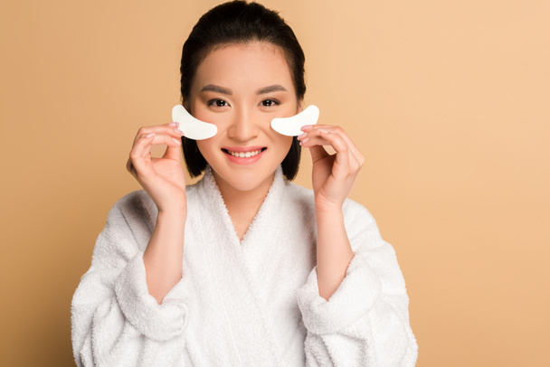 smiling beautiful asian woman in bathrobe holding eye patches on beige background - Fotoğraf, Görsel