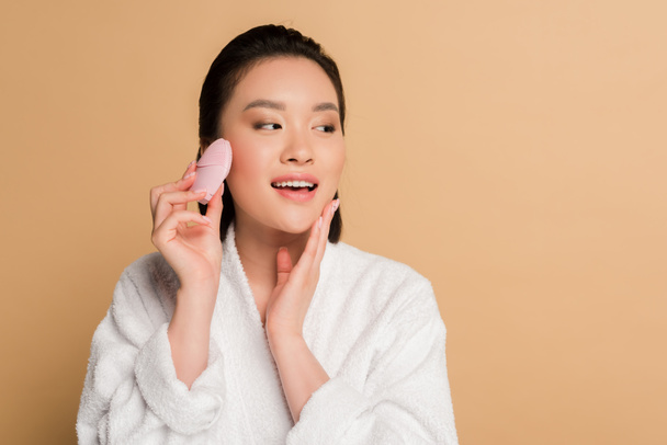 beautiful asian woman in bathrobe using facial cleansing brush on beige background - Фото, изображение