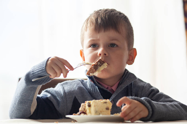 Leuke kleine jongen in café eet een groot stuk taart met een vork. Desserts voor kinderen. Hij geniet ervan.. - Foto, afbeelding