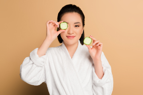smiling beautiful asian woman in bathrobe with cucumber slices on beige background - Photo, Image
