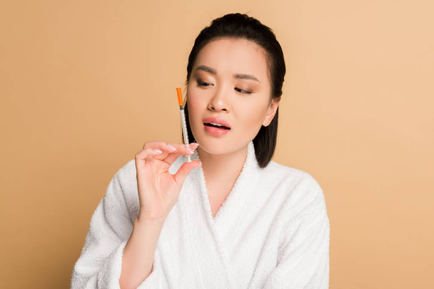 beautiful asian woman in bathrobe looking at syringe for beauty injection on beige background - Fotó, kép