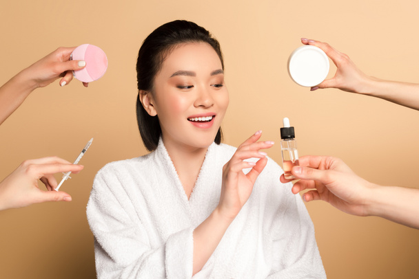 lächelnd schöne asiatische Frau im Bademantel in der Nähe der Hände mit Gesichtsreinigungsbürste, Spritze, Kosmetikcreme und Öl auf beigem Hintergrund - Foto, Bild
