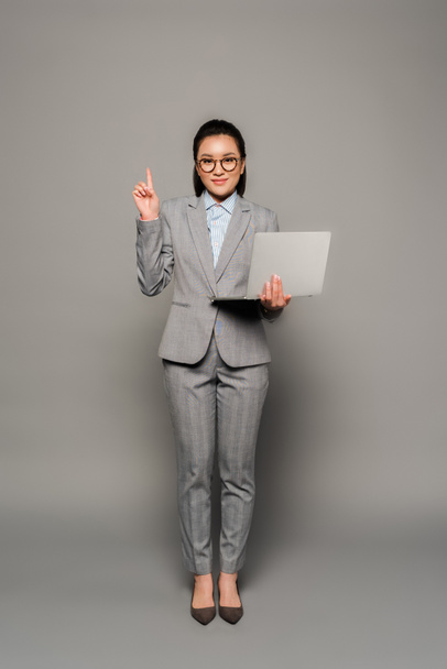 uśmiechnięta młoda bizneswoman w okularach za pomocą laptopa i pokazując gest idei na szarym tle - Zdjęcie, obraz