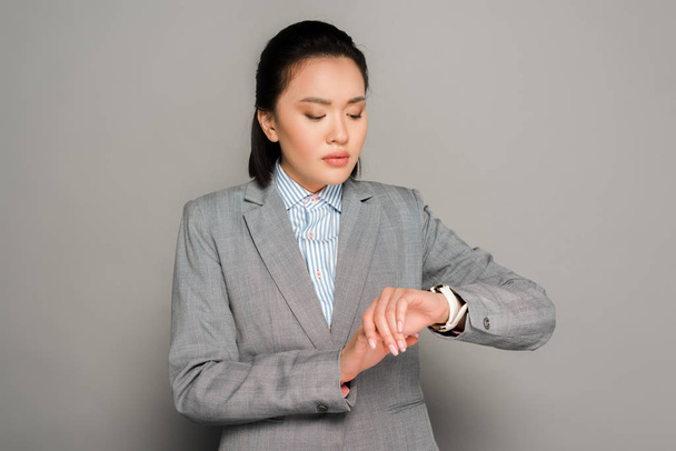 νεαρή επιχειρηματίας με κοστούμι κοιτάζοντας ρολόι χειρός σε γκρι φόντο - Φωτογραφία, εικόνα