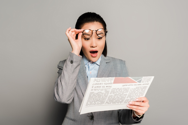 schockierte junge Geschäftsfrau mit Brille liest Zeitung auf grauem Hintergrund - Foto, Bild